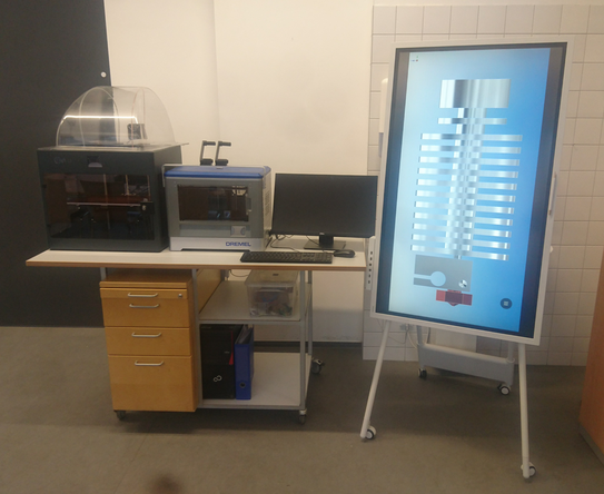 Arbeitsplatz additive Fertigung Das Bild zeigt einen Hochwagen der als Arbeitsplatz für additive Fertigung ausgerüstet ist. Auf der Arbeitsfläche stehen zwei 3d Drucker und ein Rechnerarbeitsplatz. Rechts neben dem Hochwagen steht ein großer fahrbarer Monitor der einen Druckkopf eines 3d Druckers zeigt.