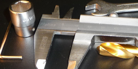 Werkzeuge eines Mechanikers Das Bild zeigt einen Maulschlüssel, eine Messschieber und eine golden beschichteten Spiralbohrer in Anschnitt auf einer dunklen Unterlage.