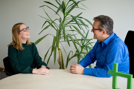 Das Bild zeigt zwei Mitarbeiter der TU Dortmund sitzend an einem Konferenztisch. Die Mitarbeiter führen ein Gespräch.