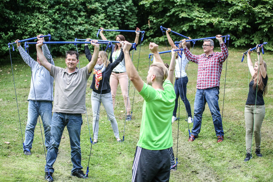 Auf dem Bild ist eine Gruppe von Menschen zu sehen die Gymnastik mit Gummibändern macht, die sie über ihre Köpfe halten.
