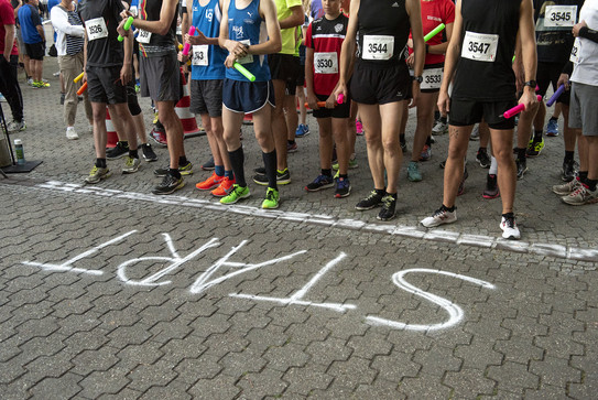 Das Bild zeigt die mit Kreide auf Asphalt gemalte Startlinie des Campuslaufs in Dortmund. Hinter der Starrtlinie sieht man die Beine und Oberkörper der auf den Start wartenden Teilnehmer.